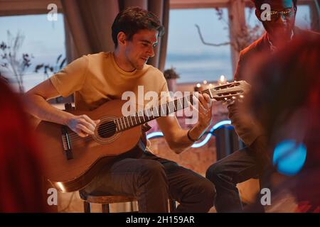 Young man sitting on chair and playing guitar for his friends in the room Stock Photo
