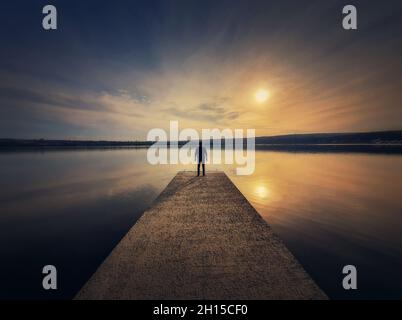 Man standing alone on the pier facing the sunset reflected in the calm lake water. Scenic view with a lonely person on the dock looking at the horizon Stock Photo