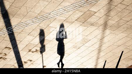 Tactile floor paving indicators and shadow silhouette of a young woman walking alone on city street square in sepia black and white Stock Photo