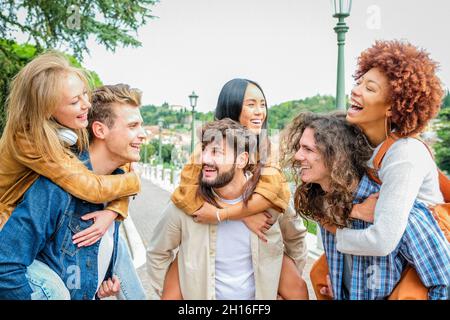 Group of friends having fun walking on city street at holiday  - Six happy young multi-ethnic men giving their girlfriends piggyback ride