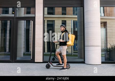 Smiling messneger man standing outdoors with electric push scooter. Delivery man with termal backpack in the city. Stock Photo