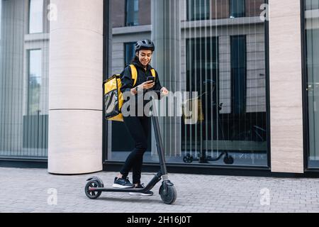 Delivery woman using smartphone while standing in the city with electric push scooter Stock Photo