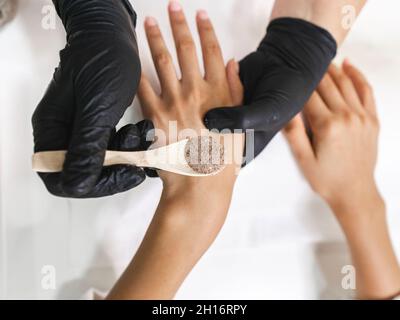 Top view of crop unrecognizable manicurist in gloves with wooden spoon with scrub above hands of client at table in salon Stock Photo