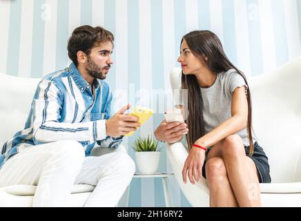 Young lady and bearded male sitting on armchairs against wall while browsing cellphones and looking at each other Stock Photo