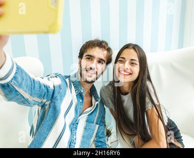 Young couple sitting on white armchairs and smiling while taking selfie on smartphone Stock Photo