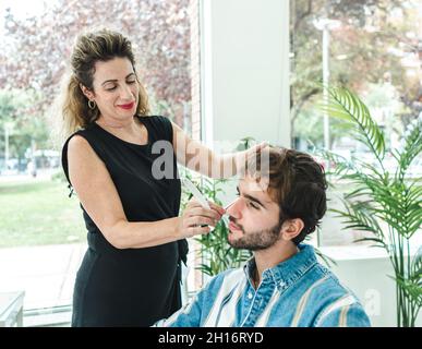 Content female master in black clothes cutting male client in barbershop against window Stock Photo