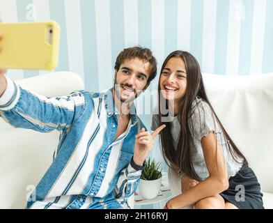 Young couple sitting on white armchairs and smiling while taking selfie on smartphone Stock Photo