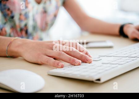 Crop anonymous female worker using keyboard while sitting at table near computer in light workspace Stock Photo