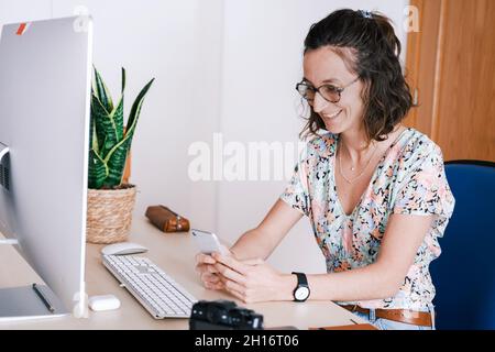 Glad female in casual outfit and eyeglasses sitting at desk with computer and browsing smartphone Stock Photo