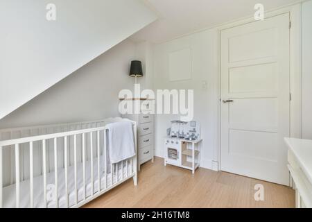 White crib with towel placed in light stylish bedroom with lamp on chest of drawers and toy near closed door Stock Photo