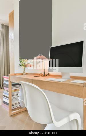 Modern computer with black screen and keyboard placed on wooden desk with lamp and flowers vase in light workroom with minimalist interior Stock Photo