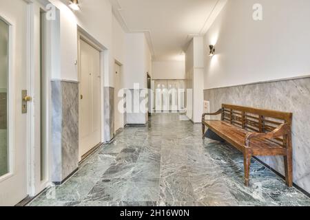 Aged shabby wooden bench placed in hallway of spacious building with marble tiled walls and white doors Stock Photo