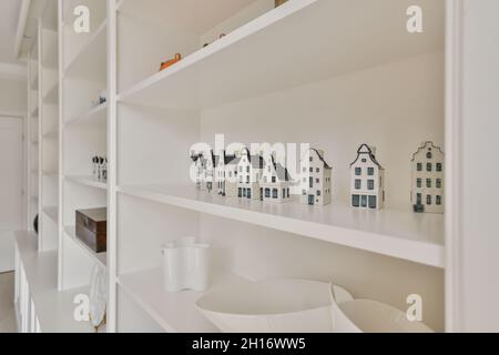 Small ceramic houses arranged in row on shelf of white bookcase in light room Stock Photo