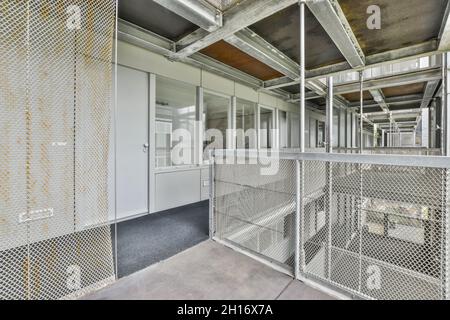 Narrow hall with door and windows against rusty metal fences under roof with beams in daytime Stock Photo