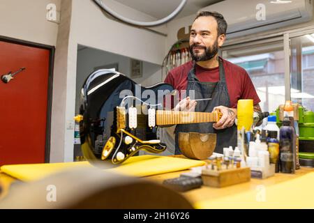 Happy bearded craftsman in apron standing at table and fixing electric guitar while working in professional workshop Stock Photo