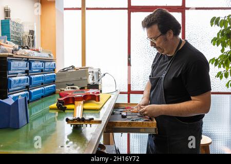 Concentrated adult male master in apron and eyeglasses standing near table with various tools while preparing to repair electric guitar in workshop ne Stock Photo