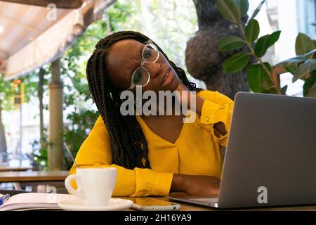 Thoughtful African American female freelancer in eyeglasses looking away while sitting at table with netbook and cup of hot drink in cafeteria Stock Photo