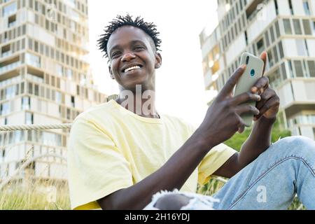 Smiling young black male wearing light yellow t shirt and blue jeans and sneakers sitting on grass and browsing smartphone Stock Photo