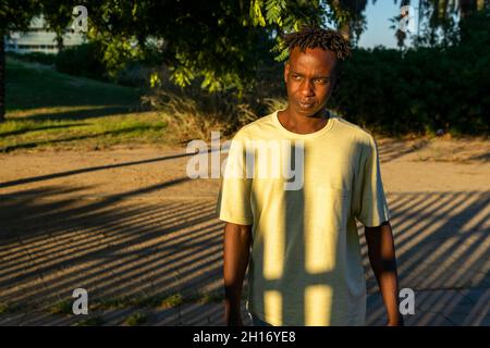 Young African American male in casual clothes standing on alley in green park and looking away thoughtfully in sunset light Stock Photo