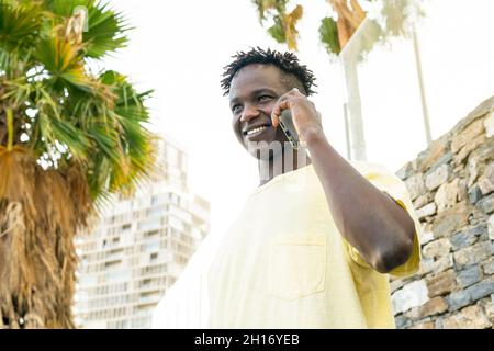 African American male with short dreadlocks hair wearing casual t shirt talking on mobile phone near brick wall in town Stock Photo