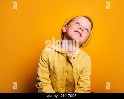 Happy boy in vivid yellow raincoat jacket and beanie hat laughing and looking away against yellow background in studio Stock Photo