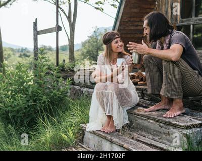 Barefoot hippie couple sharing a coffee while talking and looking at each other on a wooden porch Stock Photo