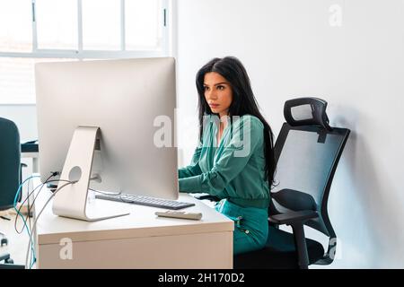 Side view of concentrated female worker browsing internet on computer while sitting at desktop on white background in office during work Stock Photo