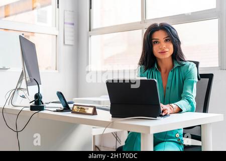Elegant female employee in formal wear looking at camera while sitting on white background at desk with modern computer during work Stock Photo