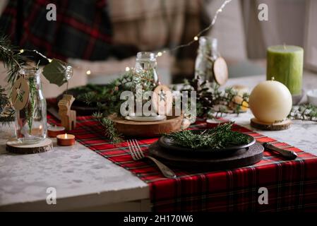 Christmas table setting with wreath on the plate, decorative wooden ornaments and red checkered tablecloth with yellow lights on the background Stock Photo
