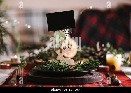 Christmas table setting with reindeer of wood with blank name card on ceramic plate on red checkered tablecloth on the background Stock Photo