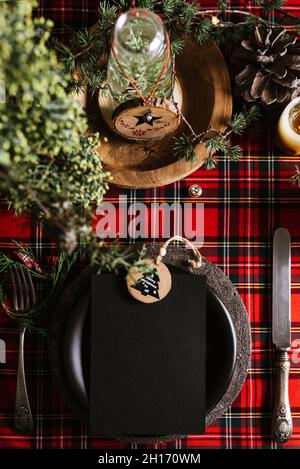 Holiday place setting with empty menu and wooden ornament, on checkered red tablecloth, on the christmas table, defocused lights. Stock Photo