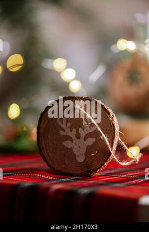 Round wooden ornament with the drawing of a reindeer, on the red checkered tablecloth and defocused lights, Christmas background. Stock Photo