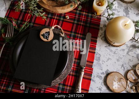 Top view of christmas table setting with empty menu on ceramic plate and red checkered tablecloth with yellow lights on the background Stock Photo
