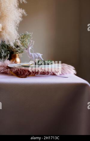 From above christmas table settings with a wreath decoration and white and golden dinnerware and white reindeer on fluffy placemat on pink tablecloth Stock Photo