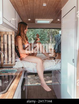 Side view of cheerful female traveler sitting on bed with dog in van and playing ukulele during trip in Spain Stock Photo