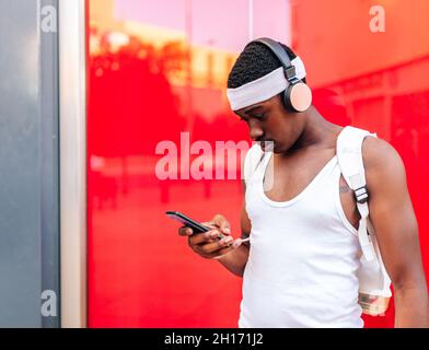 Focused African American male in white clothes listening to music in wireless headphones and messaging on mobile phone while standing against red wall Stock Photo