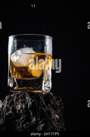 Whiskey drops falling on ice cubes served in crystal glass placed on rough surface against black background Stock Photo