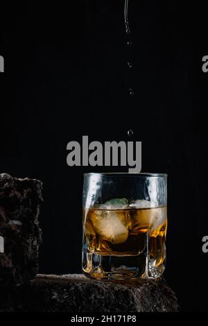 Whiskey drops falling on ice cubes served in crystal glass placed on rough surface against black background Stock Photo
