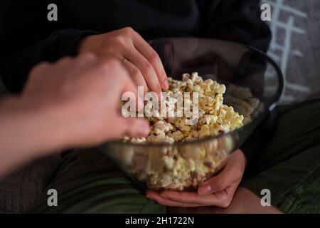 Cropped unrecognizable couple wearing casual clothes sitting on couch and eating popcorn at home Stock Photo