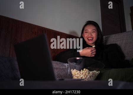 Young cheerful Asian female wearing casual clothes sitting on couch and eating popcorn at home watching movie on laptop Stock Photo