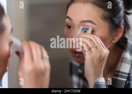 Charming concentrated Asian female using curler on eyelashes while doing makeup in front of mirror at home Stock Photo