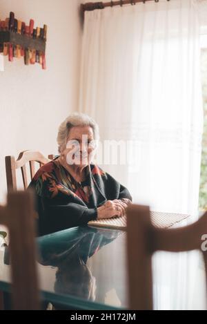 Elderly female with gray hair sitting at table in room with large window covered with tulle and smiling while looking at camera in daylight Stock Photo