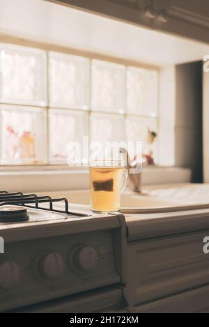 Glass cup with brewed green tea in bag placed on edge of gas stove in kitchen Stock Photo