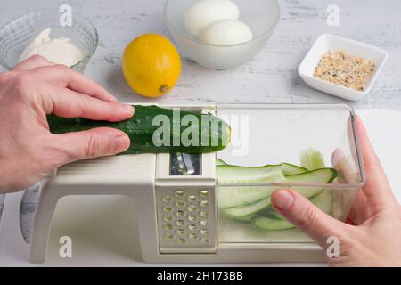 POV shot of crop faceless cook cutting fresh cucumber on slices on cutter while preparing food at table in kitchen at home Stock Photo