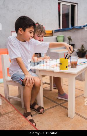 Positive kids with paintbrushes painting with colorful watercolors on paper at table with with supplies in light room with whiteboard Stock Photo
