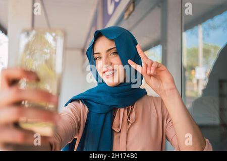 Cheerful Arab female in traditional hijab standing making peace gesture in city street and taking self shot on mobile phone Stock Photo