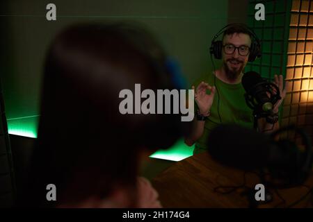 Concentrated young male and female colleagues in casual clothes and headphones sitting at table with microphones and communicating while recording pod Stock Photo