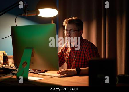 Concentrated male in checkered shirt and eyeglasses working on computer sitting at table with lamp and microphone during recording podcast Stock Photo