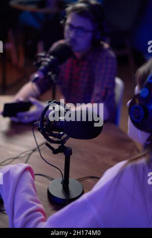 Concentrated young male and female colleagues in casual clothes and headphones sitting at table with microphones and communicating while recording pod Stock Photo