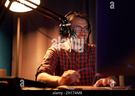 Side view of focused young male in checkered shirt and eyeglasses using computer and speaking in mic while recording podcast in dark studio Stock Photo